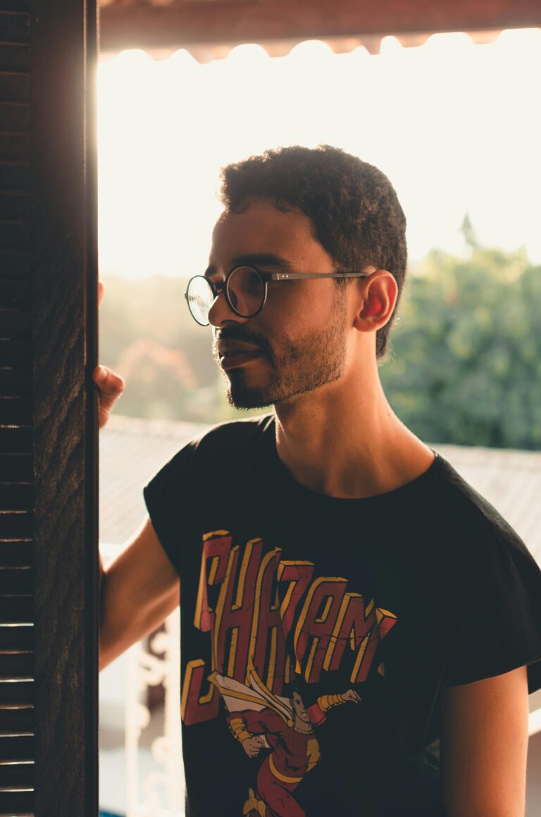 Trendy man wearing glasses and a graphic tee stands in warm sunset lighting.